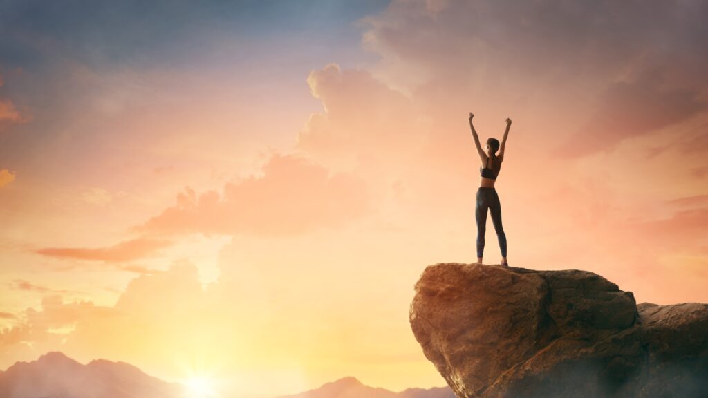 Woman standing in a triumphant mountain pose with raised arms on a mountain stop over looking a sunset. 