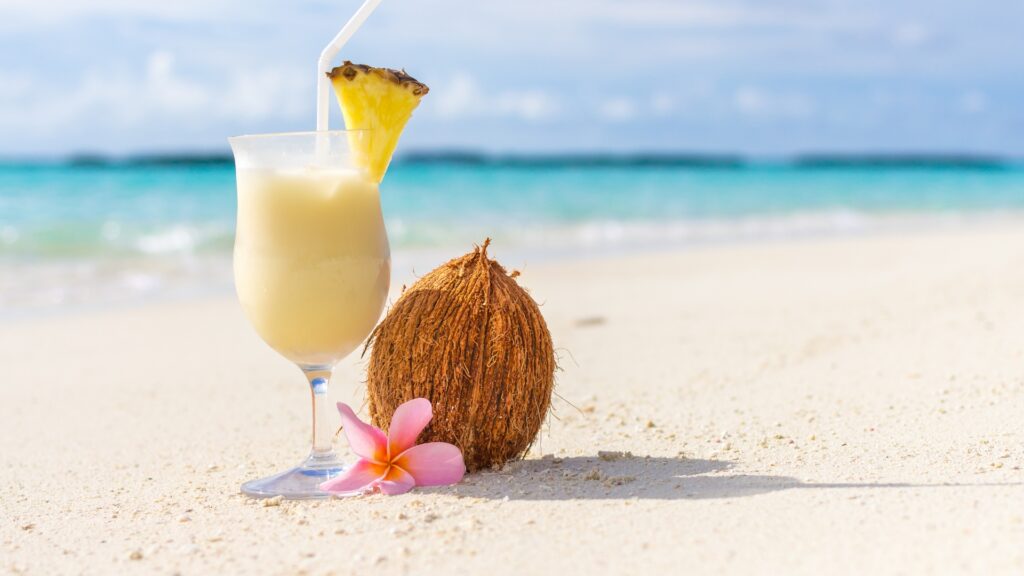 Curved glass filled with a pina colada on a white sand beach next to a coconut, pink flower and garnished with a slice of pineapple. 