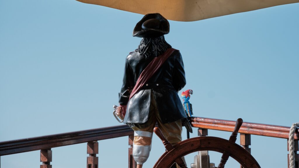Statue of a classic peg-legged pirate standing at the head of a ship looking out into the ocean with a scarlet macaw at his side.