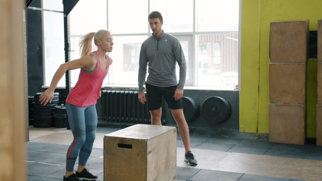 Woman jumping onto box with trainer watching fitness trend wellness trend gym box jump